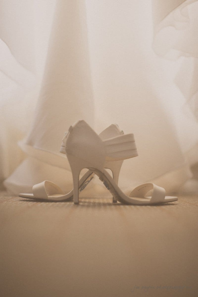 A soft-focus image of white high-heeled shoes placed on a carpet with a flowing white dress hanging behind them, suggesting a bridal setting. The lighting creates a warm, dreamy atmosphere.