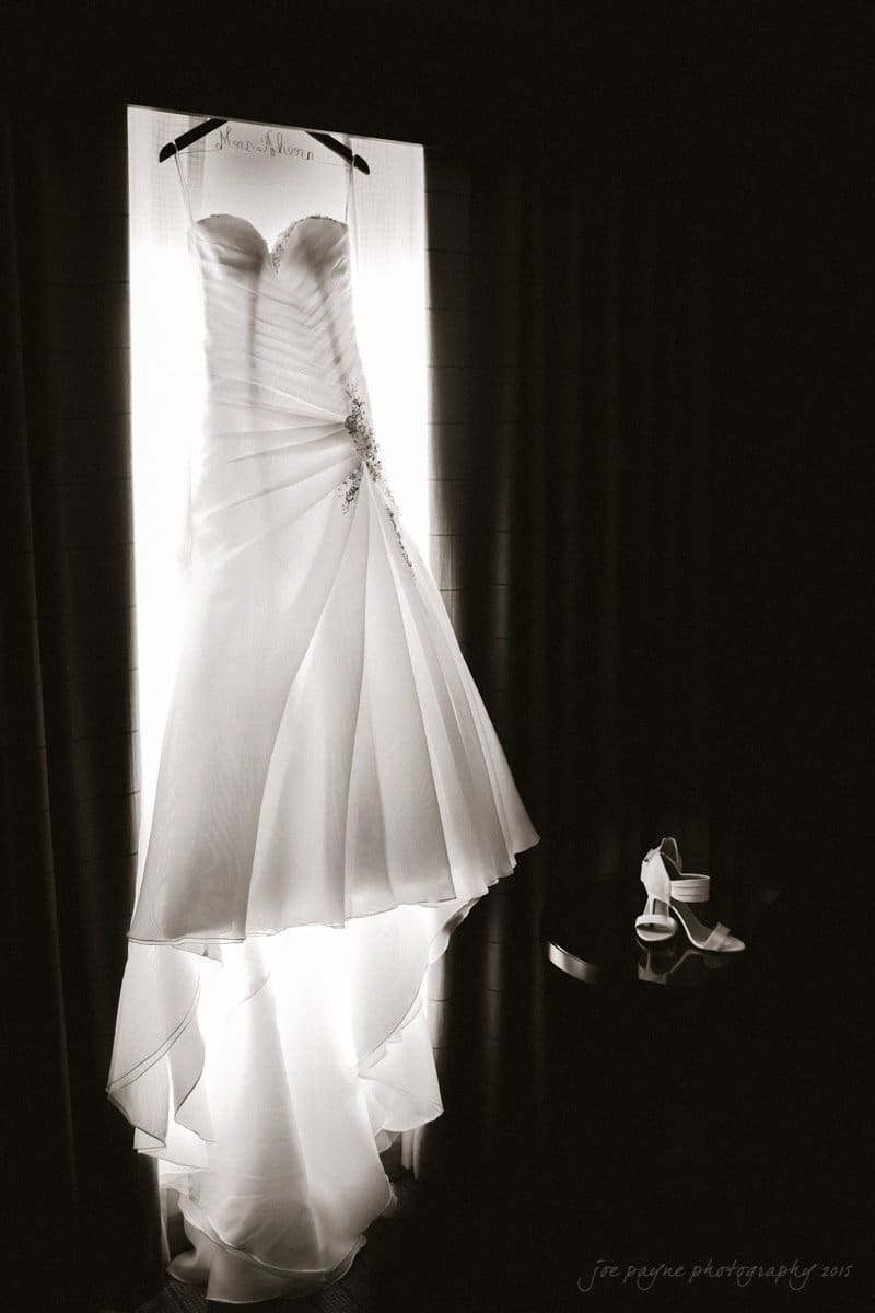 A strapless wedding dress with elegant pleats and sparkling embellishments hangs against a softly lit background. A pair of open-toe heels is placed on the right, adding to the bridal ensemble.