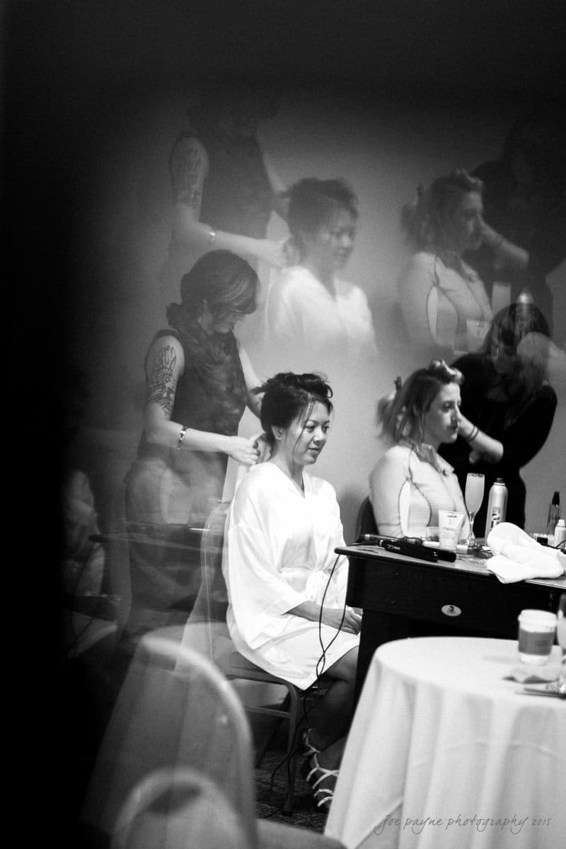 Black and white image of a woman sitting and having her hair styled. Reflections of other women also getting hairstyles are visible. The setting appears to be a preparation room, with a table in the foreground.