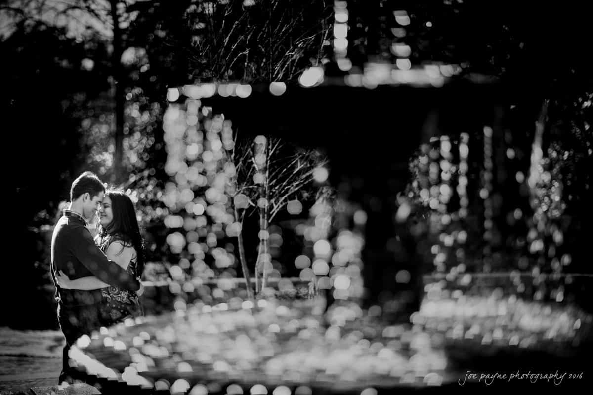 duke gardens engagement photography - yasmin & mike A black and white photo of a couple embracing near a fountain. Sunlight filters through the water droplets, creating a sparkling effect. The couple stands closely, sharing an intimate moment amidst the serene setting.