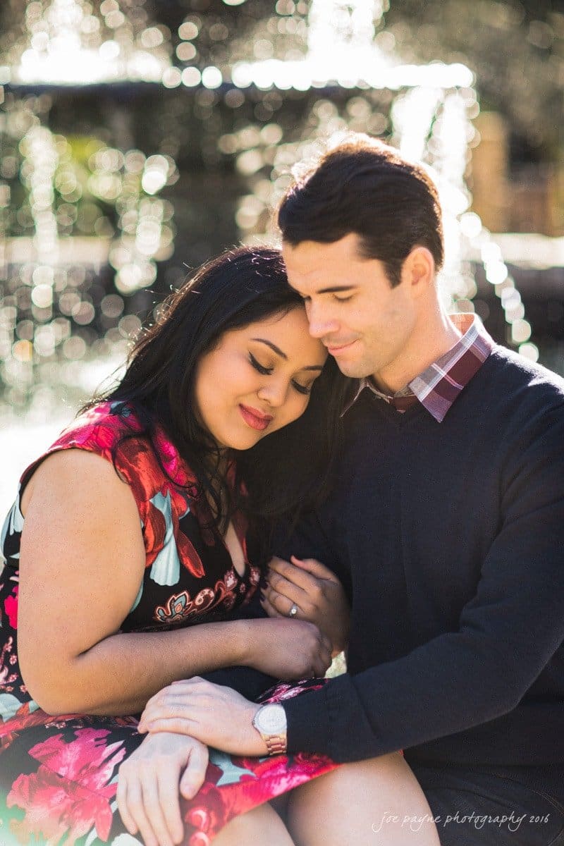 duke gardens engagement photography - yasmin & mike A couple sits closely in a sunlit outdoor setting. The woman in a floral dress leans her head on the mans shoulder, while he wears a dark sweater and holds her hand gently. Both have peaceful expressions.