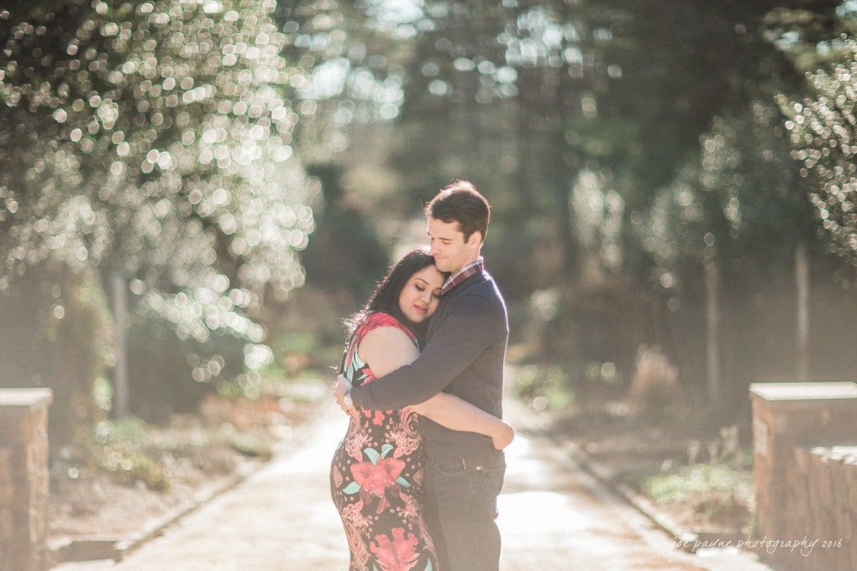 duke gardens engagement photography - yasmin & mike A couple embraces in the center of a sunlit garden path. The woman wears a floral dress, and the man is in a dark sweater. Sunlight filters through the trees, creating a dappled bokeh effect in the background.