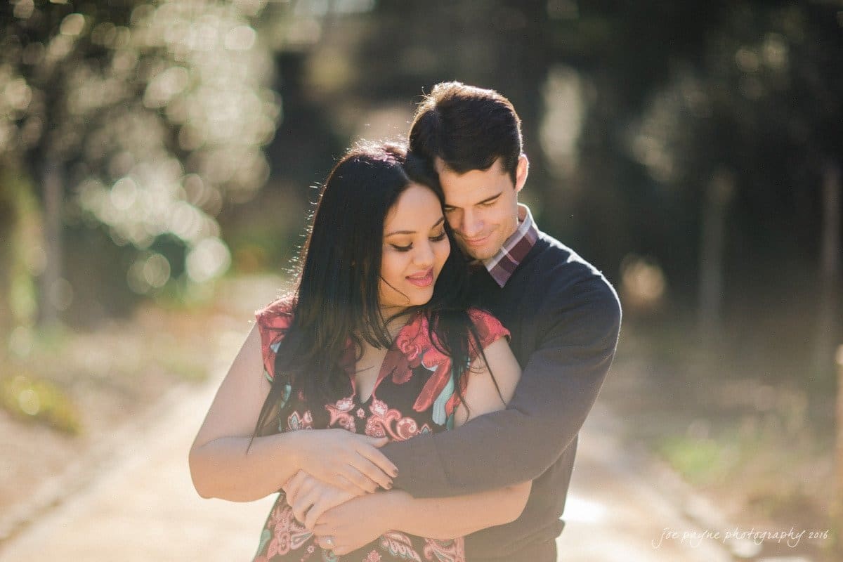 duke gardens engagement photography - yasmin & mike A couple embraces on a sunlit pathway. The woman, wearing a floral dress, stands in front, leaning back with a content smile. The man, in a dark sweater, stands behind her with his eyes closed, gently holding her. Blurred greenery surrounds them.