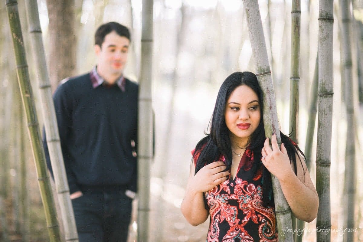 duke gardens engagement photography - yasmin & mike A woman stands among bamboo trees, looking contemplative. A man in the background gazes in her direction with hands in his pockets. The scene is softly lit, creating a serene and intimate atmosphere.