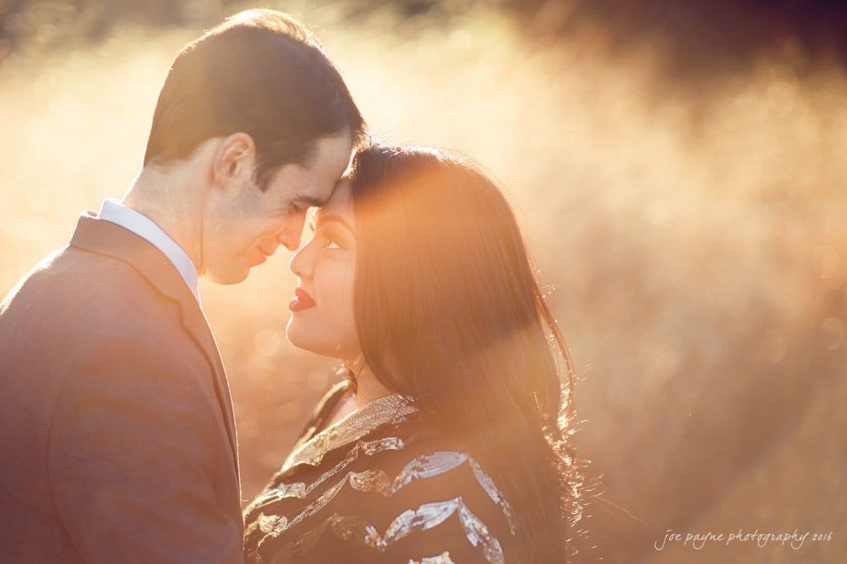 duke gardens engagement photography - yasmin & mike A couple stands close, foreheads touching and smiling warmly at each other, in a sunlit field. The man wears a suit and tie, while the woman wears a patterned dress. Sunlight creates a dreamy, glowing effect around them.