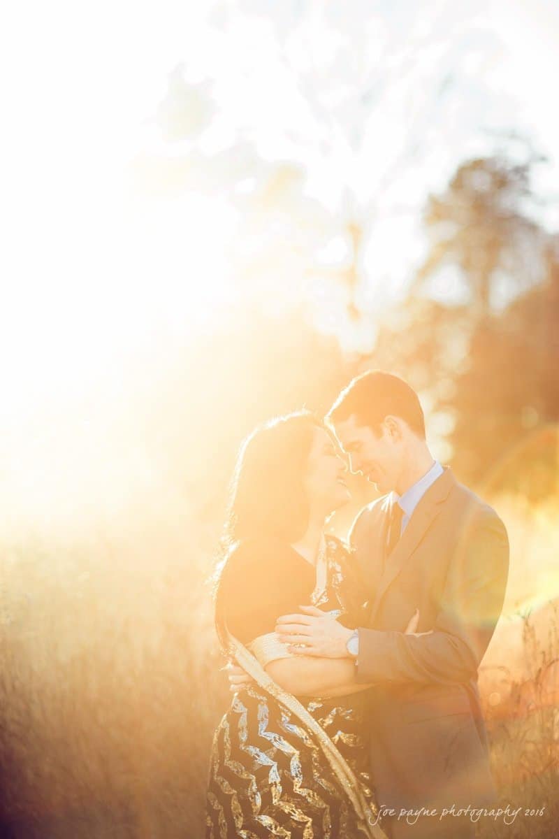 duke gardens engagement photography - yasmin & mike A couple stands embracing and smiling at each other in a sunlit field. The scene is brightly backlit, giving a warm, ethereal glow to the image. The woman wears a patterned dress, and the man is in a suit.