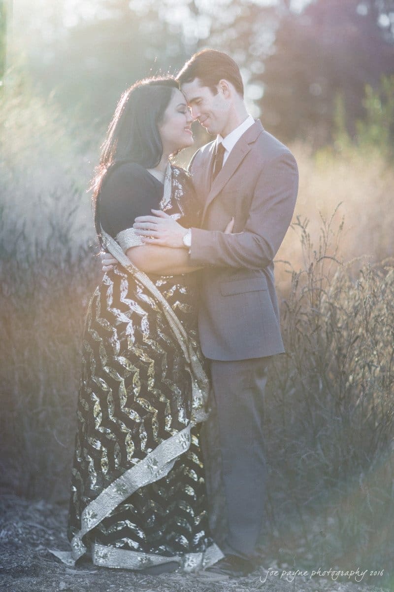 duke gardens engagement photography - yasmin & mike A couple embraces in a sunlit outdoor setting. The woman wears a black and silver sari, while the man is dressed in a gray suit. Sunlight creates a warm, glowing effect around them.