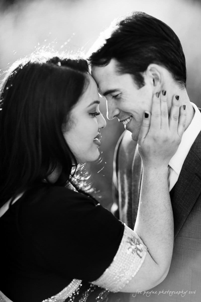 duke gardens engagement photography - yasmin & mike A black and white photo shows a couple embracing closely. The woman is gently holding the mans face, both smiling softly. They are dressed formally, and the background is softly blurred, creating an intimate atmosphere.