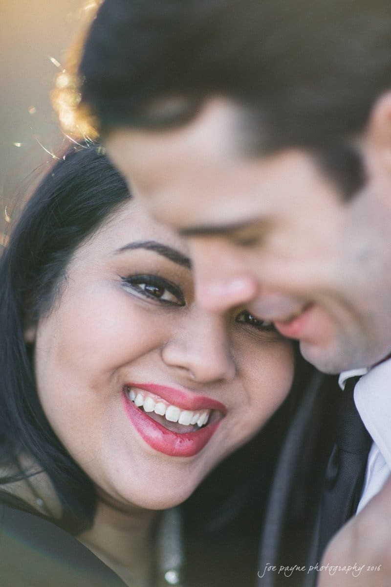 duke gardens engagement photography - yasmin & mike A close-up of a smiling couple embracing, with the woman in focus. She has dark hair and is looking at the camera, while the man is slightly blurred, leaning in, and smiling gently. The background is softly lit.