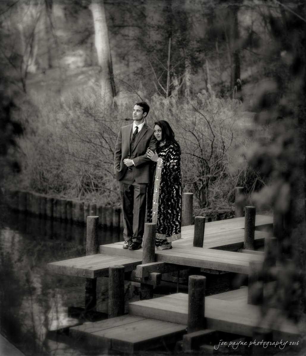 duke gardens engagement photography - yasmin & mike A couple stands together on a wooden dock by a serene lake. The man wears a suit, and the woman wears a patterned dress. They look into the distance, surrounded by trees and calm water. The image is in black and white, creating a nostalgic feel.