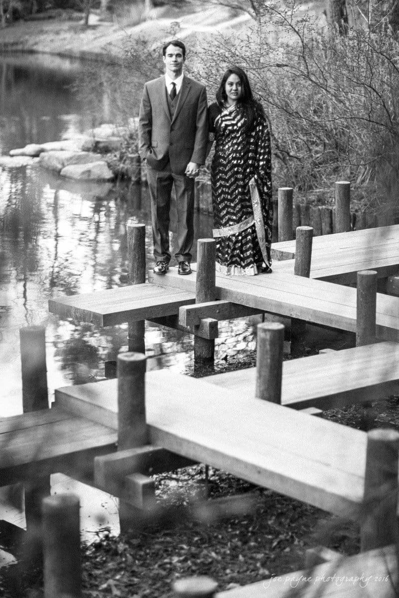 duke gardens engagement photography - yasmin & mike A couple standing on a wooden bridge over a pond, holding hands. He is in a suit, and she is in a patterned dress. The surrounding area is filled with trees and shrubs. The image is in black and white.