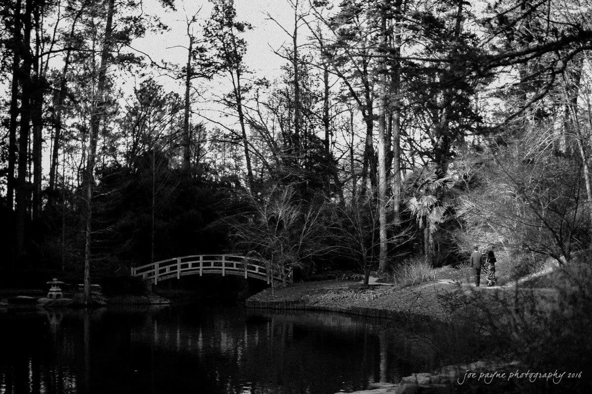 duke gardens engagement photography - yasmin & mike A monochrome image of a serene park scene with a small arched bridge over a pond, surrounded by tall trees. The reflection of the trees is visible in the water, and there are two people walking along a path on the right.