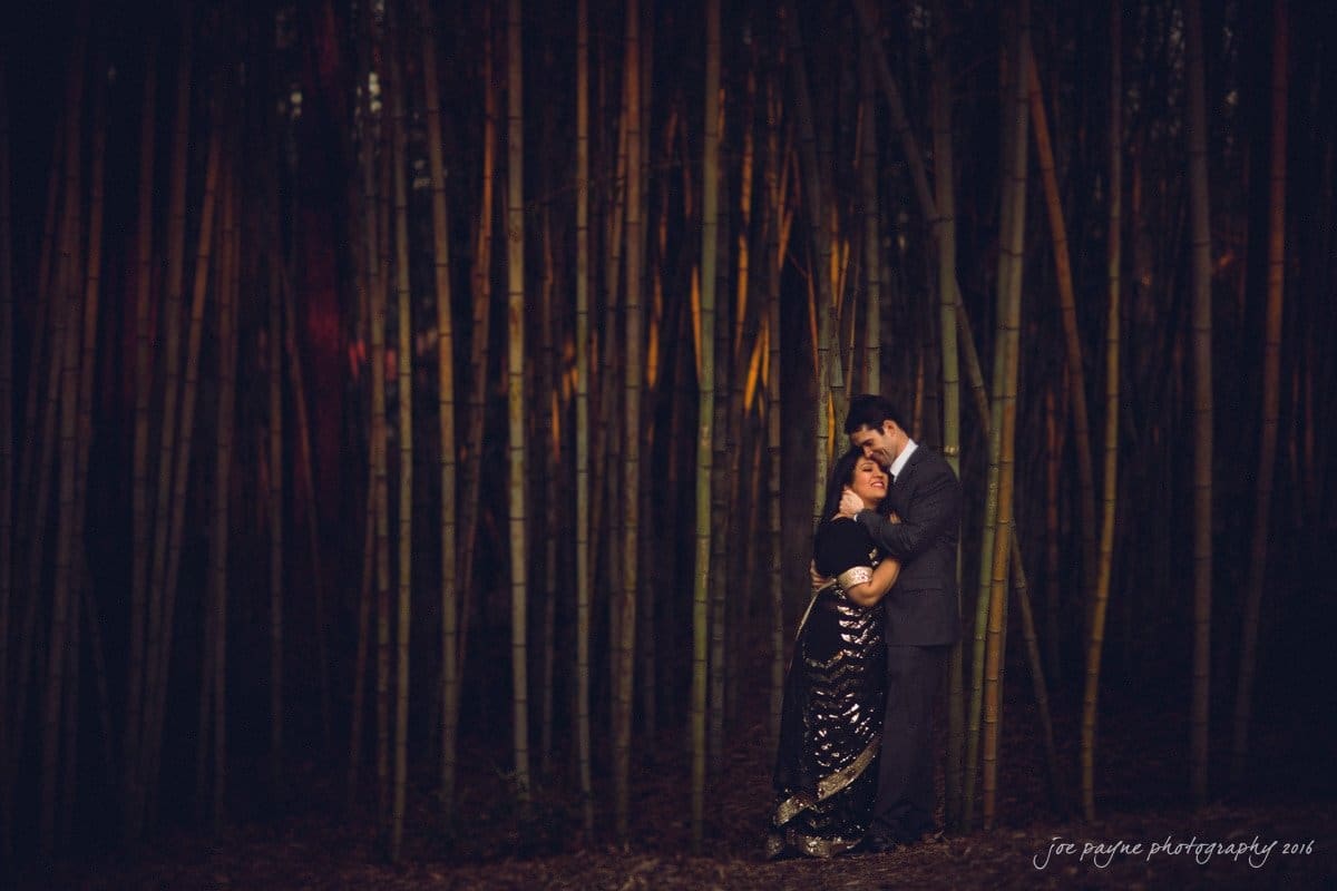 duke gardens engagement photography - yasmin & mike A couple dressed elegantly stands closely in a serene bamboo forest. The woman wears a shimmering gown and the man is in a suit. They embrace lovingly, surrounded by tall bamboo stalks and dim, atmospheric lighting.