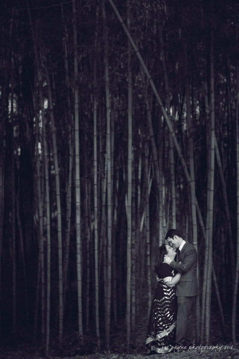 duke gardens engagement photography - yasmin & mike A couple embraces lovingly in a dense bamboo forest. They are dressed elegantly; the woman in a sequined gown and the man in a suit. The image has a monochrome effect, adding a moody, romantic atmosphere.