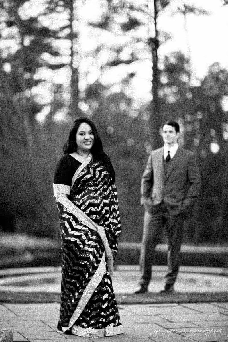 duke gardens engagement photography - yasmin & mike A woman in a patterned sari stands in the foreground smiling, while a man in a suit stands in the background. They are outdoors with trees visible in the distance. The image is in black and white.