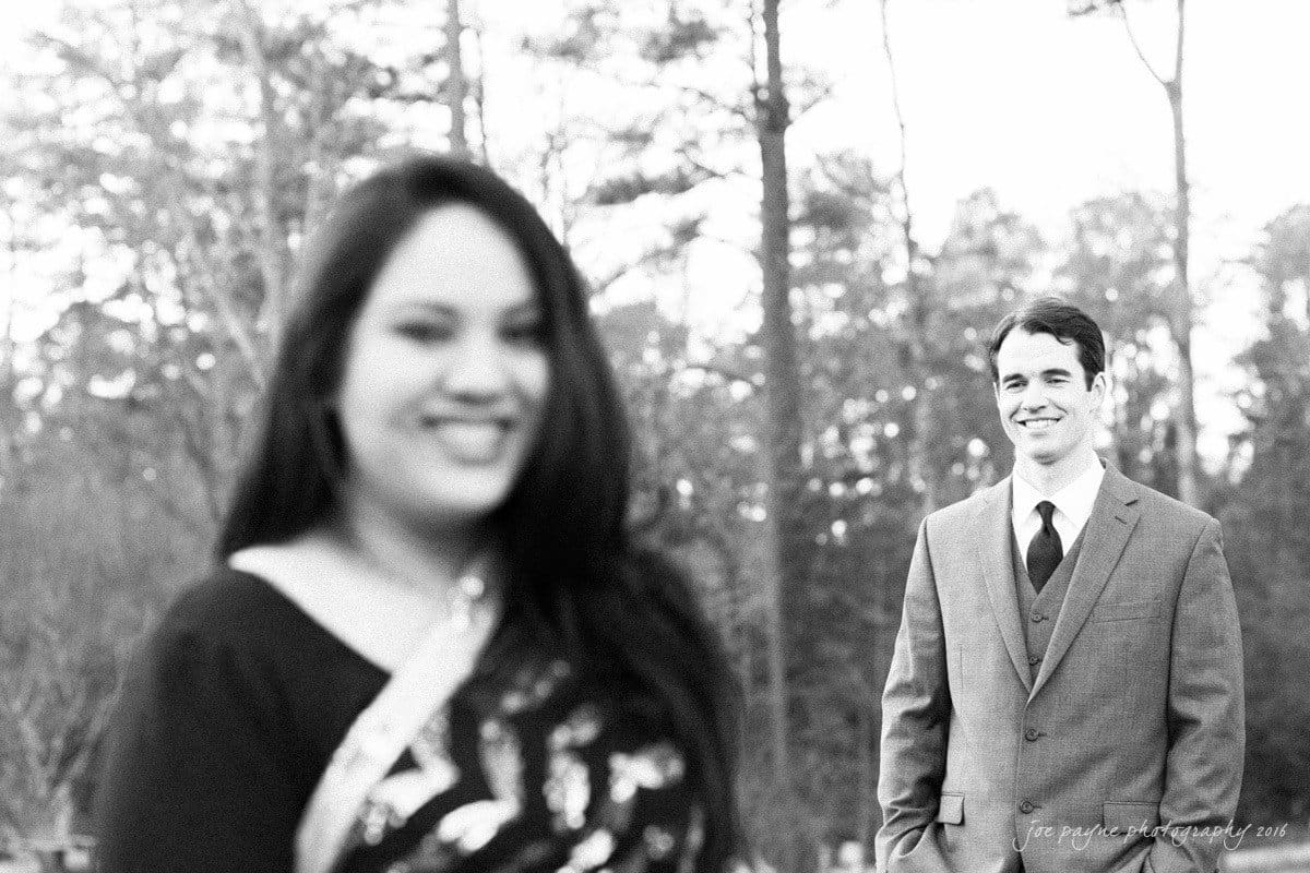 duke gardens engagement photography - yasmin & mike A woman in focus at the front, smiling, with a man in a suit blurred in the background. They are outdoors with trees surrounding them. The photo is black and white.