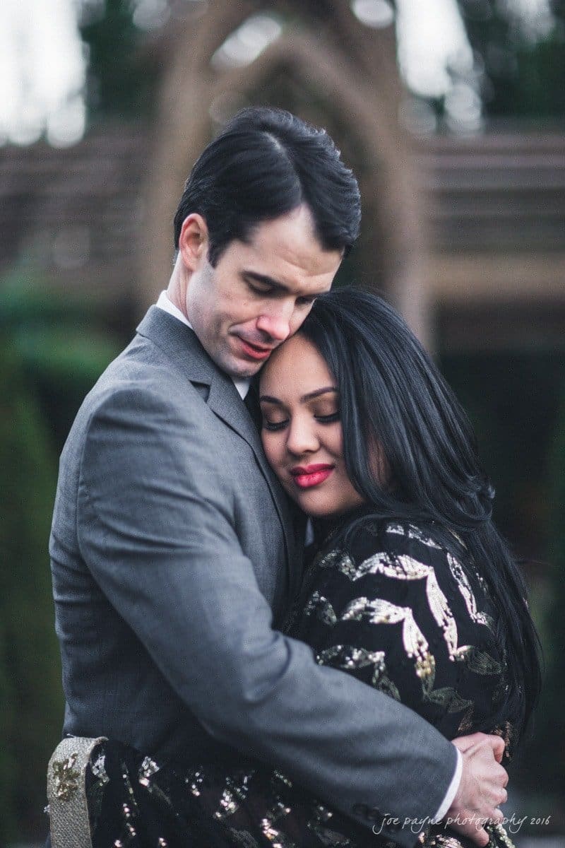 duke gardens engagement photography - yasmin & mike A couple embraces tenderly outdoors. The man in a grey suit and the woman in a black and gold dress hold each other closely, both with eyes closed. The background is blurred, suggesting a garden setting.