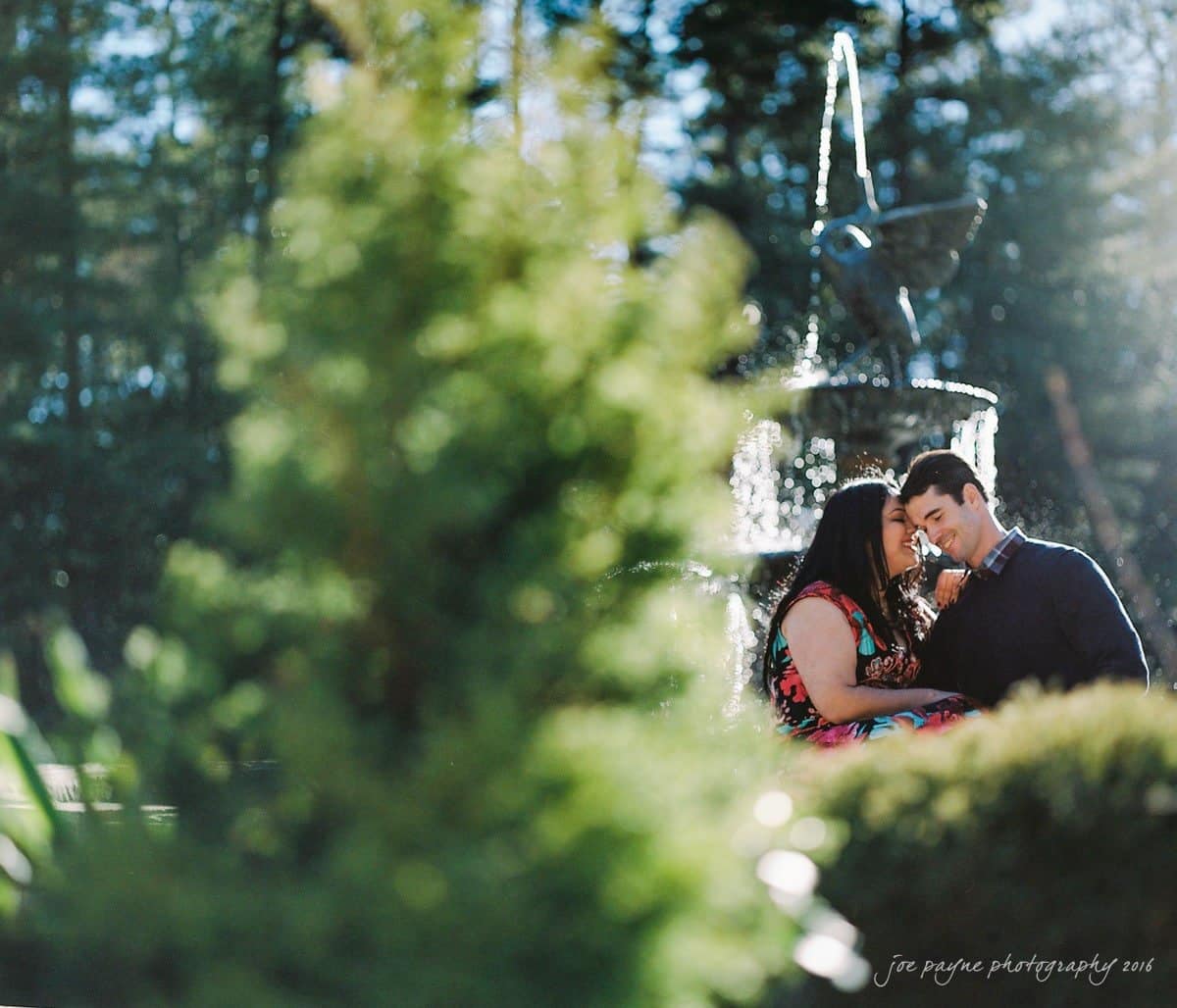duke gardens engagement photography - yasmin & mike A couple sits closely together in front of a fountain in a garden setting. The sun creates a soft glow, and greenery frames the foreground, adding a lush, serene atmosphere to the scene.