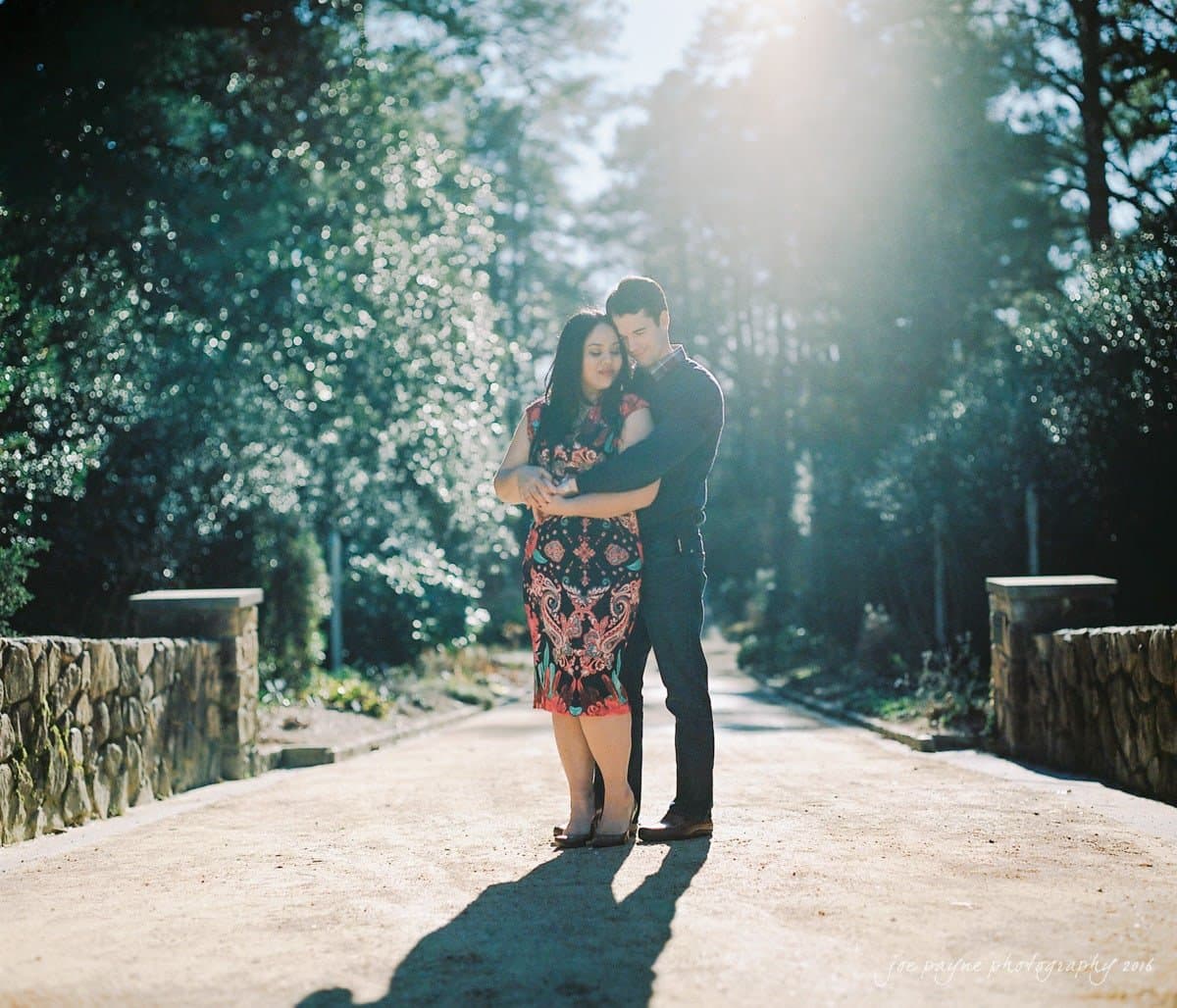 duke gardens engagement photography - yasmin & mike A couple embraces on a sunlit path surrounded by nature. The woman wears a patterned dress, while the man wears a dark outfit. Sunlight filters through the trees, casting a serene glow over the scene.