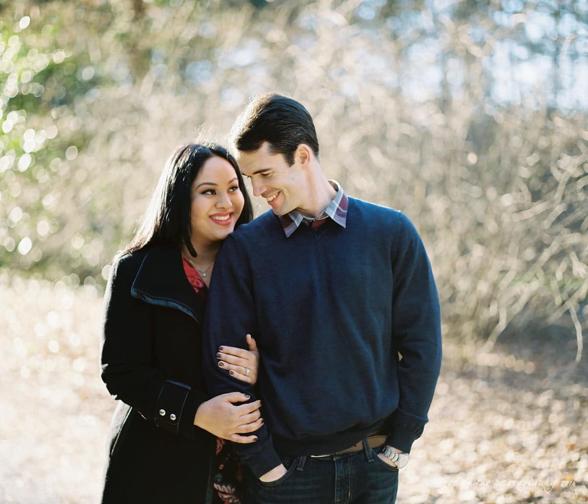 duke gardens engagement photography - yasmin & mike A couple standing together outdoors, smiling and enjoying a sunny day. The woman in a black coat holds the mans arm. The background is filled with trees and soft, natural light.