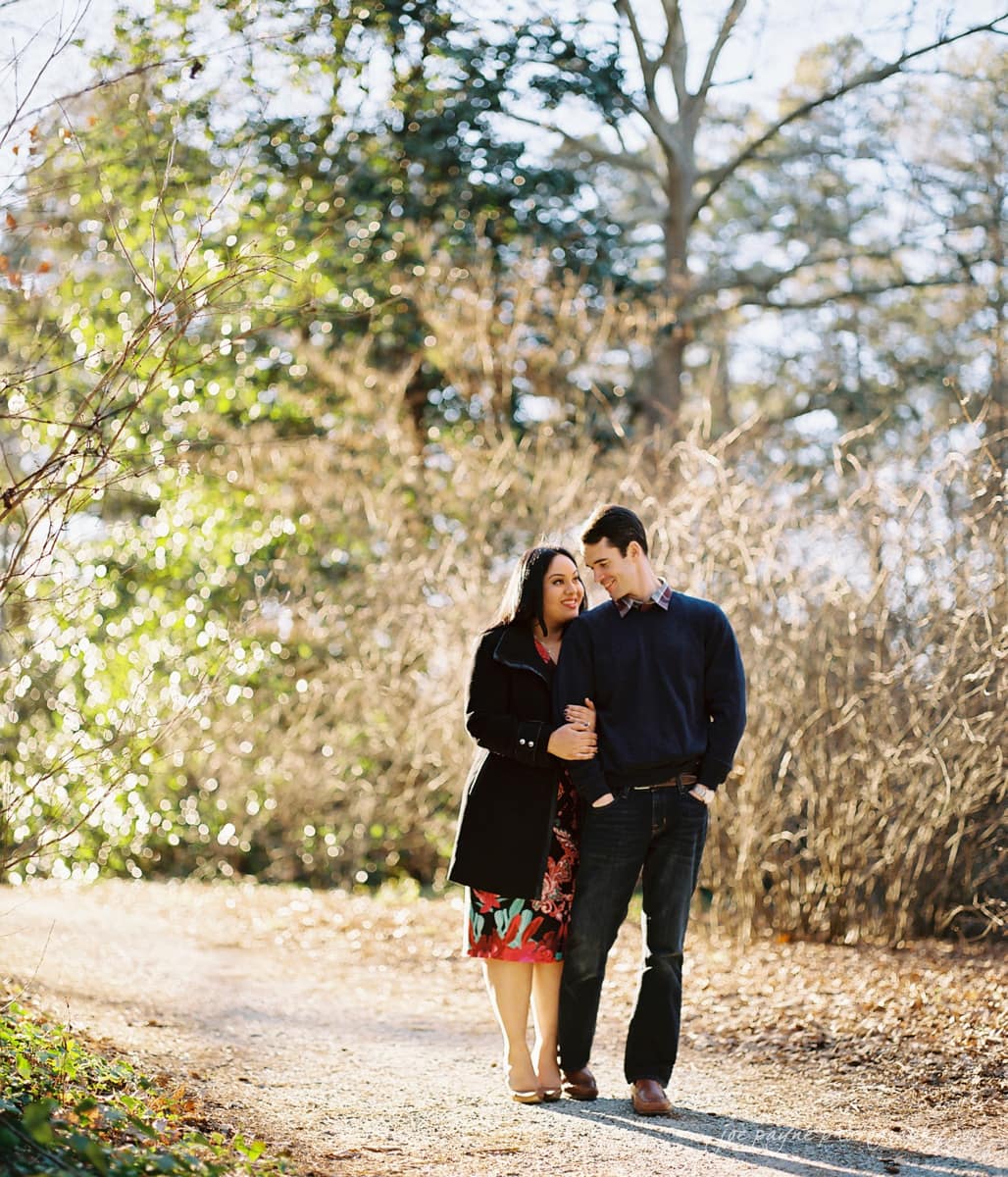 duke gardens engagement photography - yasmin & mike A couple walks arm in arm down a sunlit path surrounded by trees and dry foliage. The woman wears a floral dress and dark coat, while the man wears a blue sweater and jeans. They look at each other, smiling warmly.