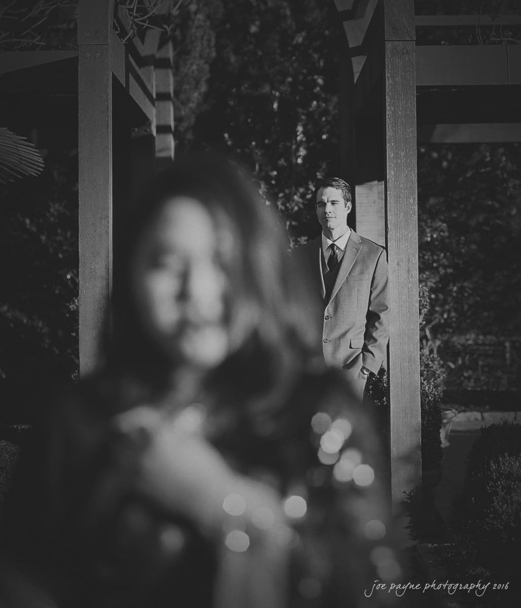 duke gardens engagement photography - yasmin & mike A black and white photo showing a man in a suit standing under a pergola in the background, while an unfocused figure is in the foreground. The scene is outdoors with plants and trees around.