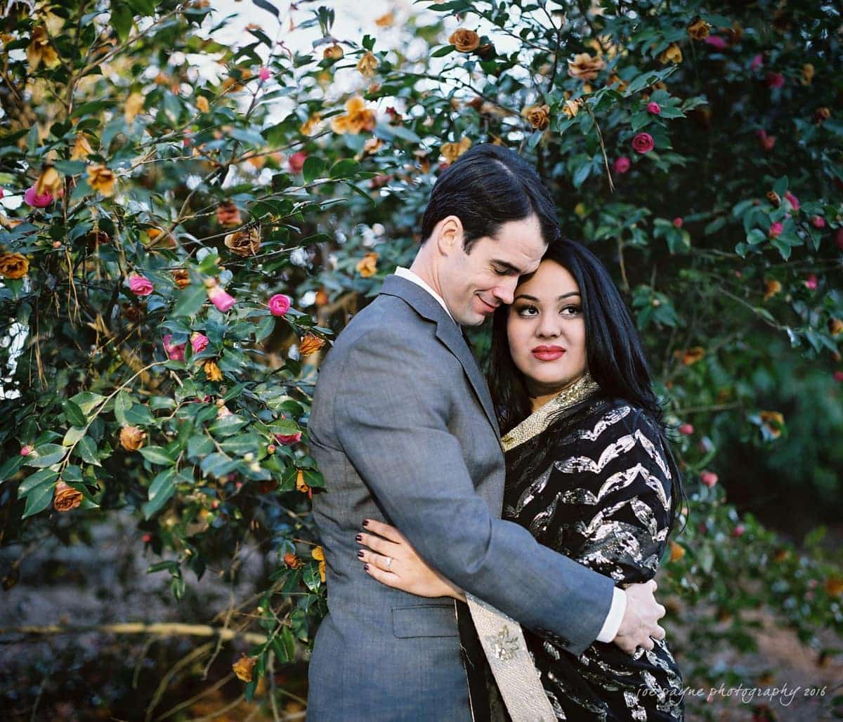 duke gardens engagement photography - yasmin & mike A couple embraces outdoors in front of a flowering bush. The man, wearing a gray suit, rests his cheek on the womans head. The woman, wearing a black and silver outfit, looks towards the camera. The bush has pink and yellow flowers.