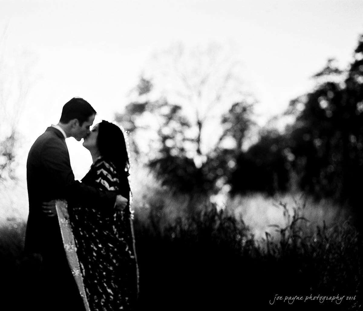 duke gardens engagement photography - yasmin & mike A couple is embracing in a grassy field, sharing a kiss. The scene is in black and white, with tall grasses and blurred trees in the background. The image conveys a romantic and serene atmosphere.
