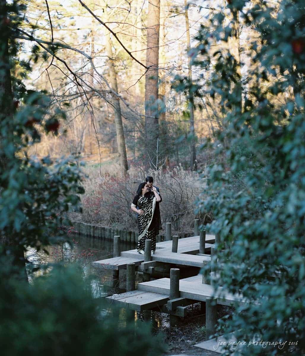 duke gardens engagement photography - yasmin & mike A couple stands romantically on a wooden dock surrounded by lush greenery and calm water. Bare trees in the background suggest an early spring or late autumn setting. The scene is serene and intimate.