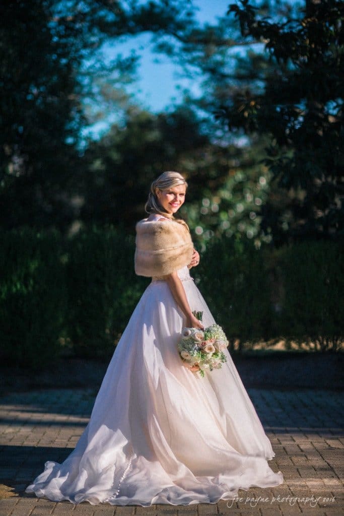 A bride in a white gown and fur shawl stands outdoors on a sunlit path, holding a bouquet of white and pink flowers. The background features greenery and trees.