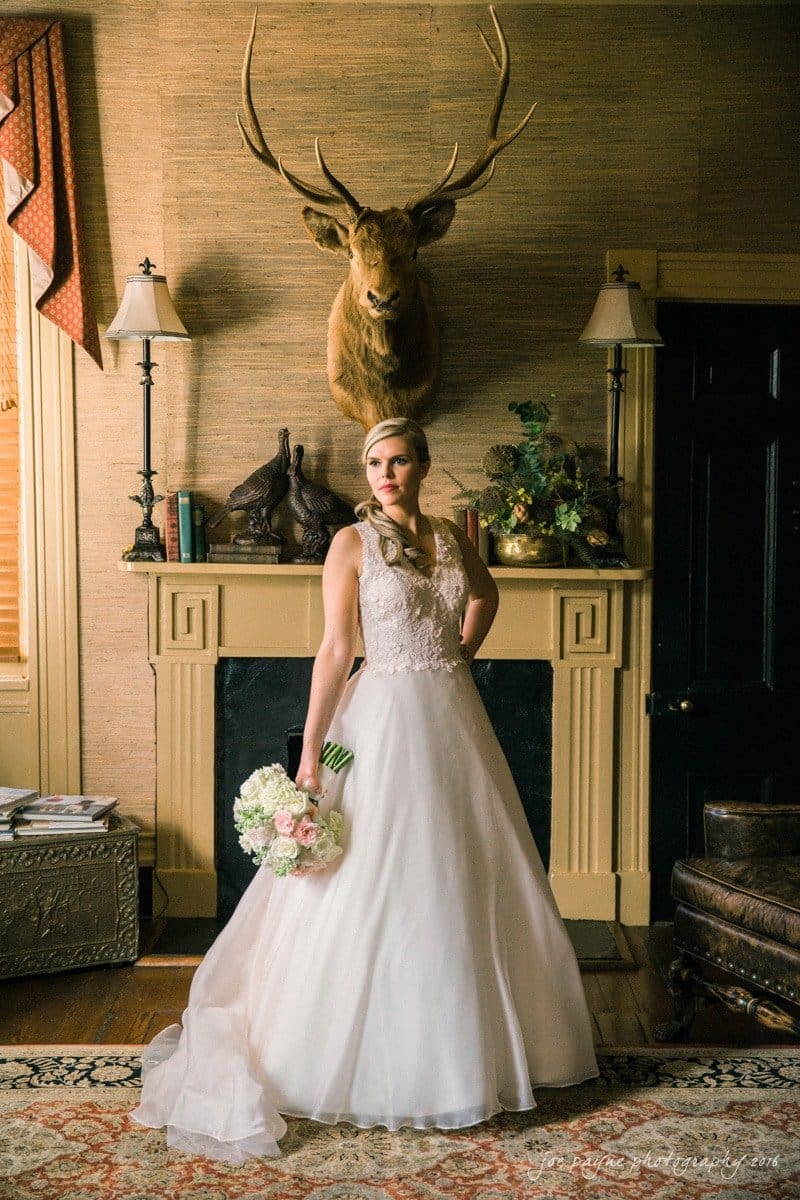 raleigh bridal photography session - jackie at the sutherland A bride in an elegant white gown holds a bouquet of flowers, standing in a warmly lit room. Behind her, theres a fireplace with a mounted deer head on the wall and bookshelves on each side.