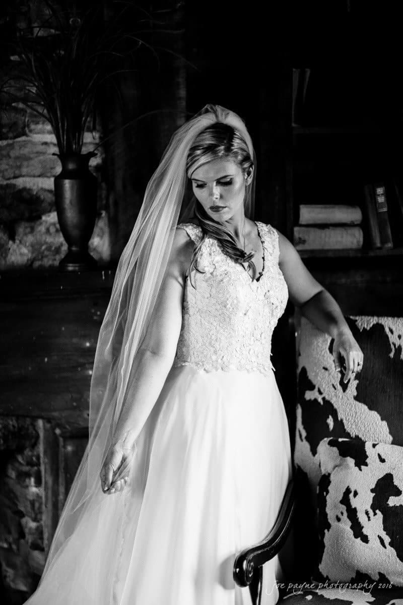 raleigh bridal photography session - jackie at the sutherland A bride in a white wedding dress and veil stands indoors with a contemplative expression. She is posing near a patterned chair and a shelf, with one arm gently outstretched. The setting has a vintage, elegant feel. Black and white photograph.