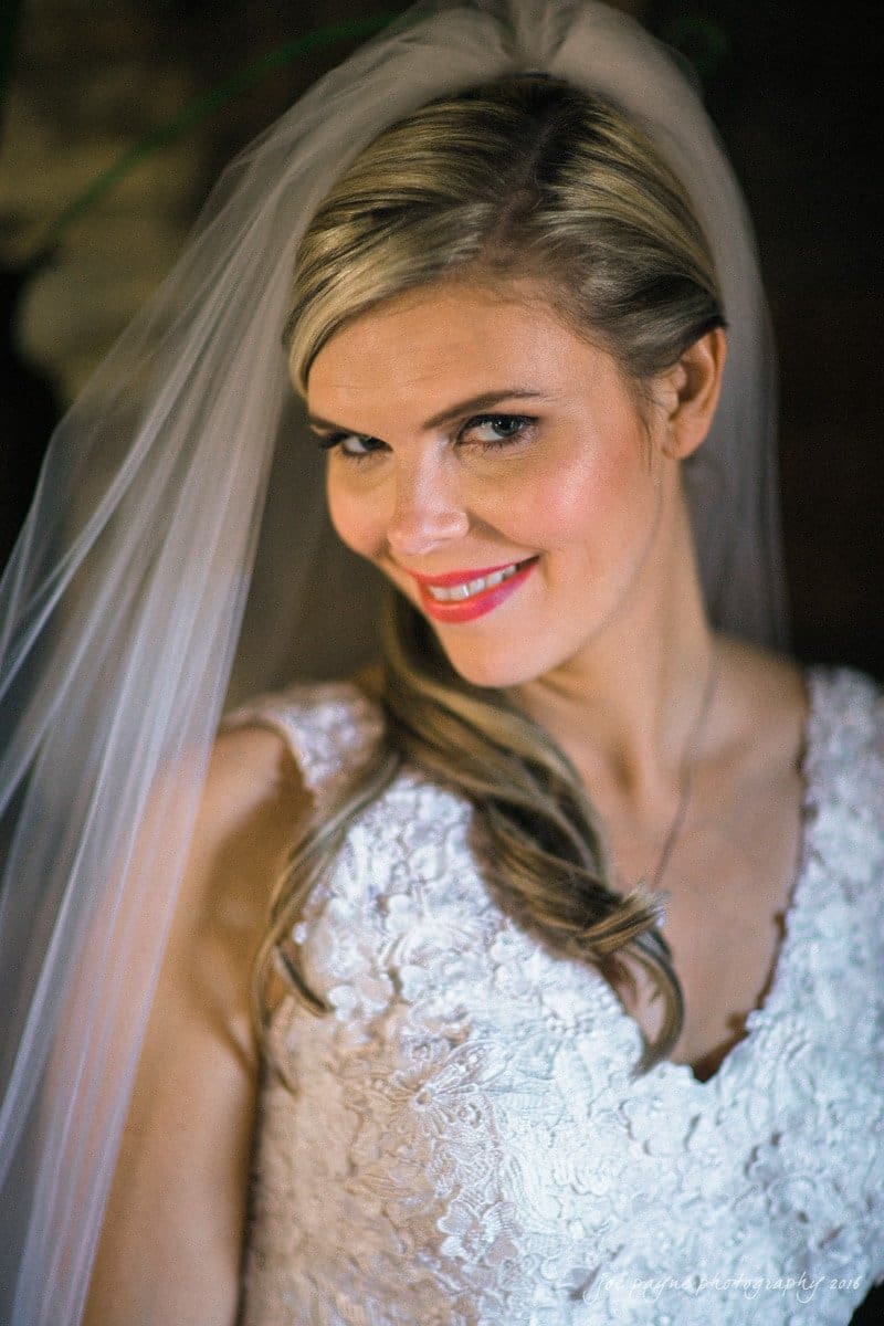 raleigh bridal photography session - jackie at the sutherland A bride with blonde hair and a side-swept hairstyle smiles at the camera. She wears a white lace wedding dress and a veil. The background is softly blurred, drawing focus to her expression.