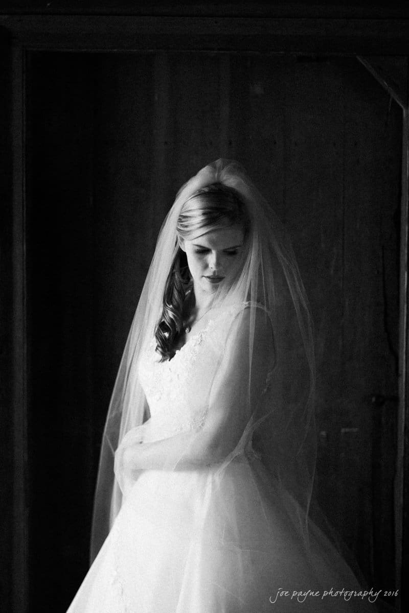 raleigh bridal photography session - jackie at the sutherland Bride in a white wedding dress and veil stands in front of a wooden background, looking down with a gentle expression. The monochrome image highlights her serene and elegant pose.