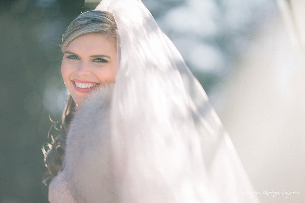 raleigh bridal photography session - jackie at the sutherland A bride with a wide smile poses outdoors. She wears a veil and a light fur stole. Soft, natural lighting creates a dreamy atmosphere, with gentle bokeh effects in the background.