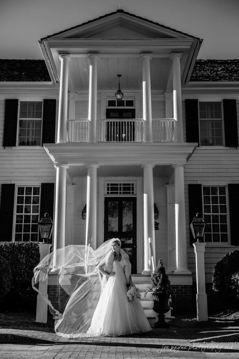 raleigh bridal photography session - jackie at the sutherland A bride in a flowing gown poses with her veil dramatically blown to the side in front of an elegant two-story building with tall columns and dark shutters. The image is in black and white, highlighting the contrast and structure.