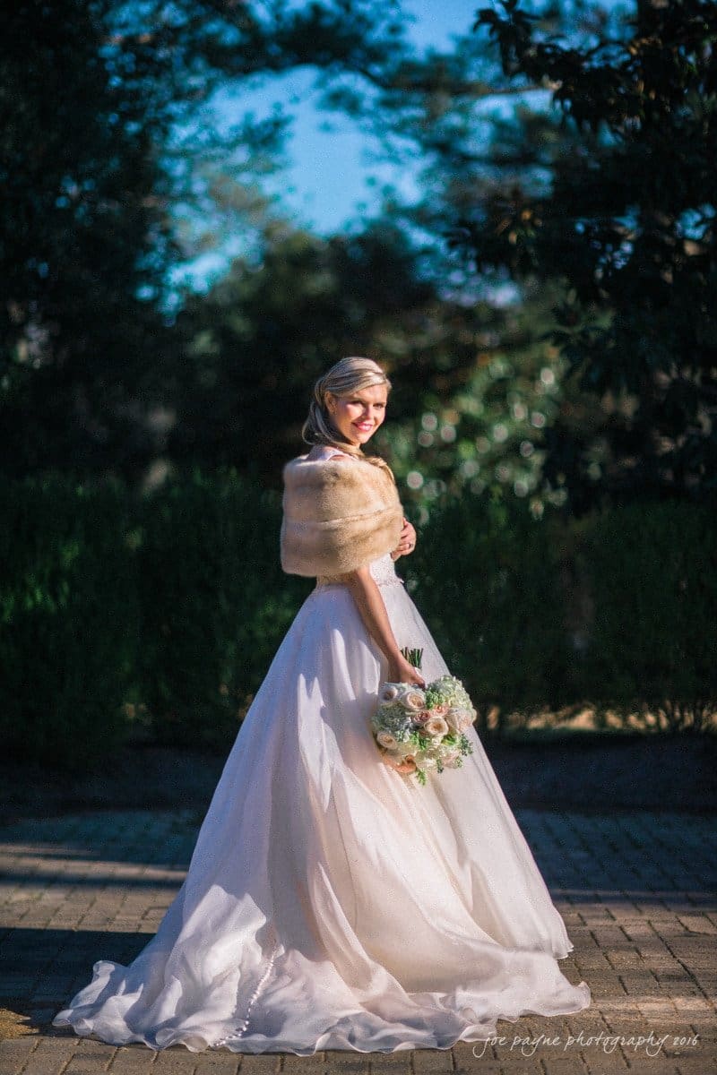 raleigh bridal photography session - jackie at the sutherland A bride in a white gown and fur shawl stands outdoors on a sunlit path, holding a bouquet of white and pink flowers. The background features greenery and trees.