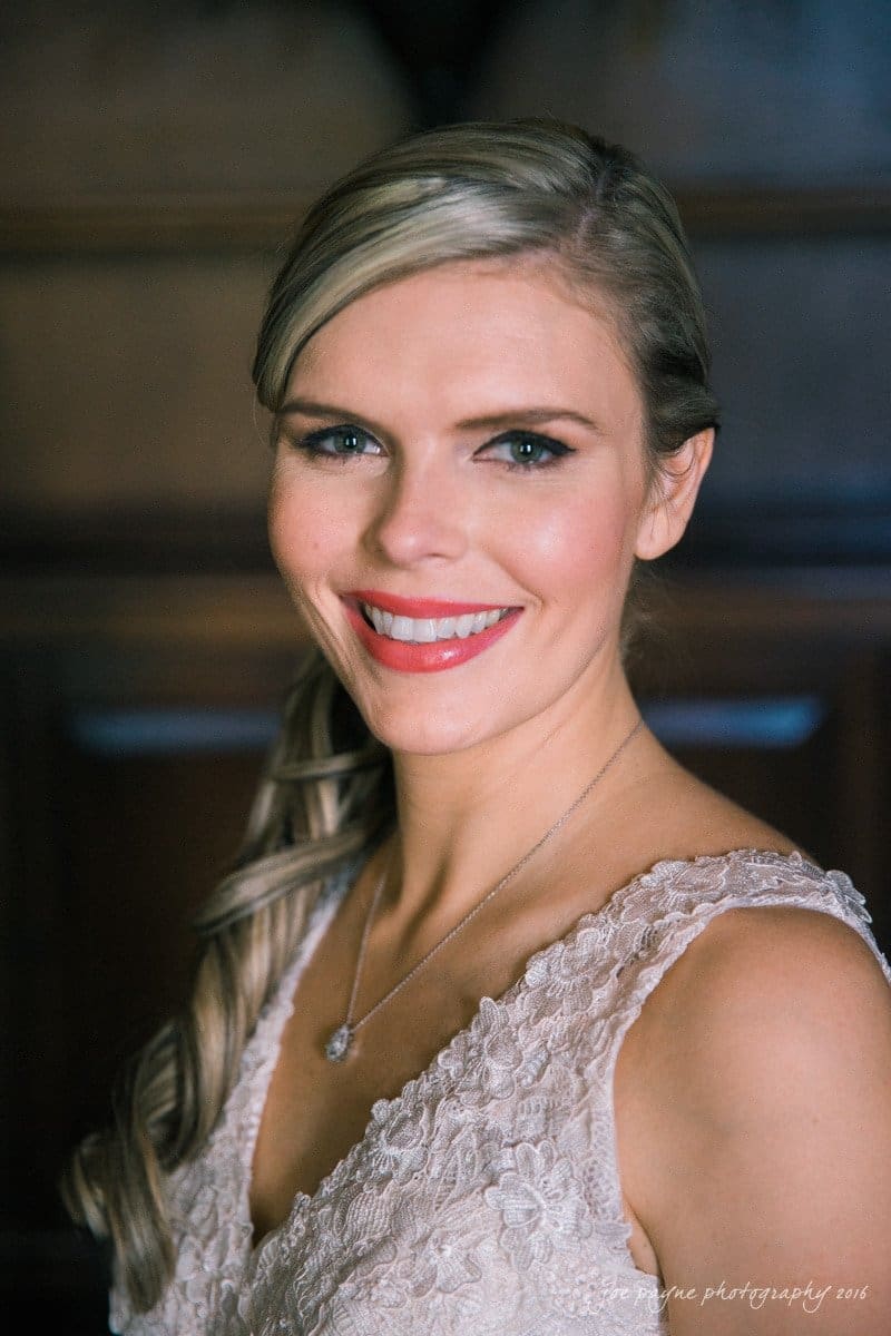 raleigh bridal photography session - jackie at the sutherland A woman with long, light blonde hair styled in a side braid smiles. She wears a sleeveless, lace dress and a necklace. The background is softly blurred, creating a warm atmosphere.