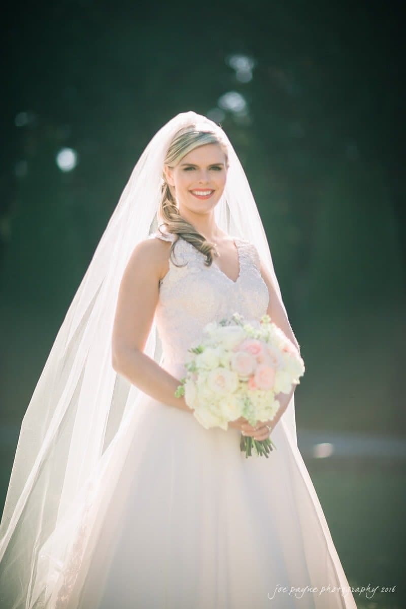 raleigh bridal photography session - jackie at the sutherland A bride in a white dress smiles, holding a bouquet of pink and white flowers. She is wearing a veil and is standing outdoors with sunlight creating a soft glow around her.
