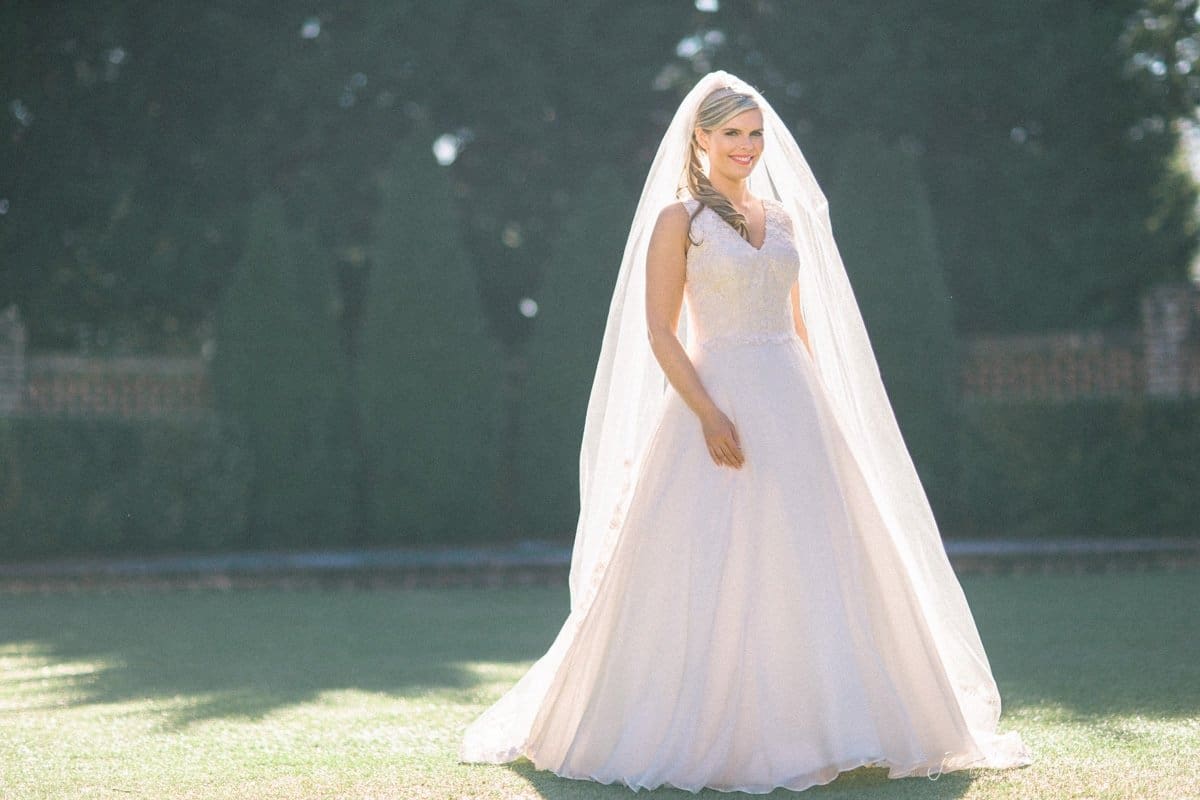 raleigh bridal photography session - jackie at the sutherland A bride stands outdoors, smiling, in a flowing white wedding dress with a long veil. The background is blurred greenery, bathed in soft sunlight, creating a serene and joyful atmosphere.