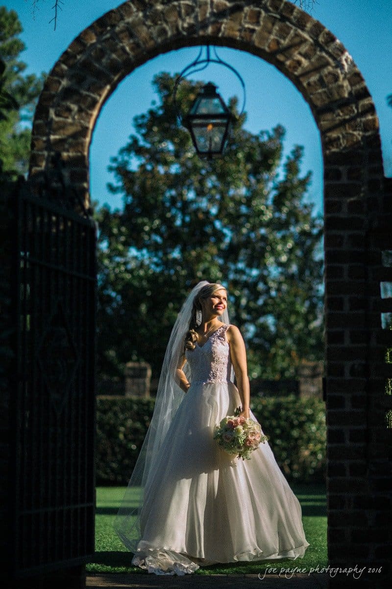 raleigh bridal photography session - jackie at the sutherland A bride stands under a brick archway, holding a bouquet and smiling. She wears a white wedding dress with lace details and a long veil. Sunlight casts a soft glow around her, highlighting her silhouette against the garden backdrop.