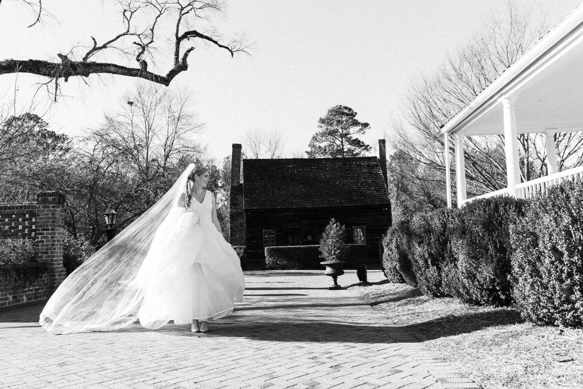 raleigh bridal photography session - jackie at the sutherland A bride in a flowing gown and veil walks along a brick path. She is outdoors, surrounded by leafless trees and shrubbery, with an old wooden building in the background. The scene is captured in black and white.