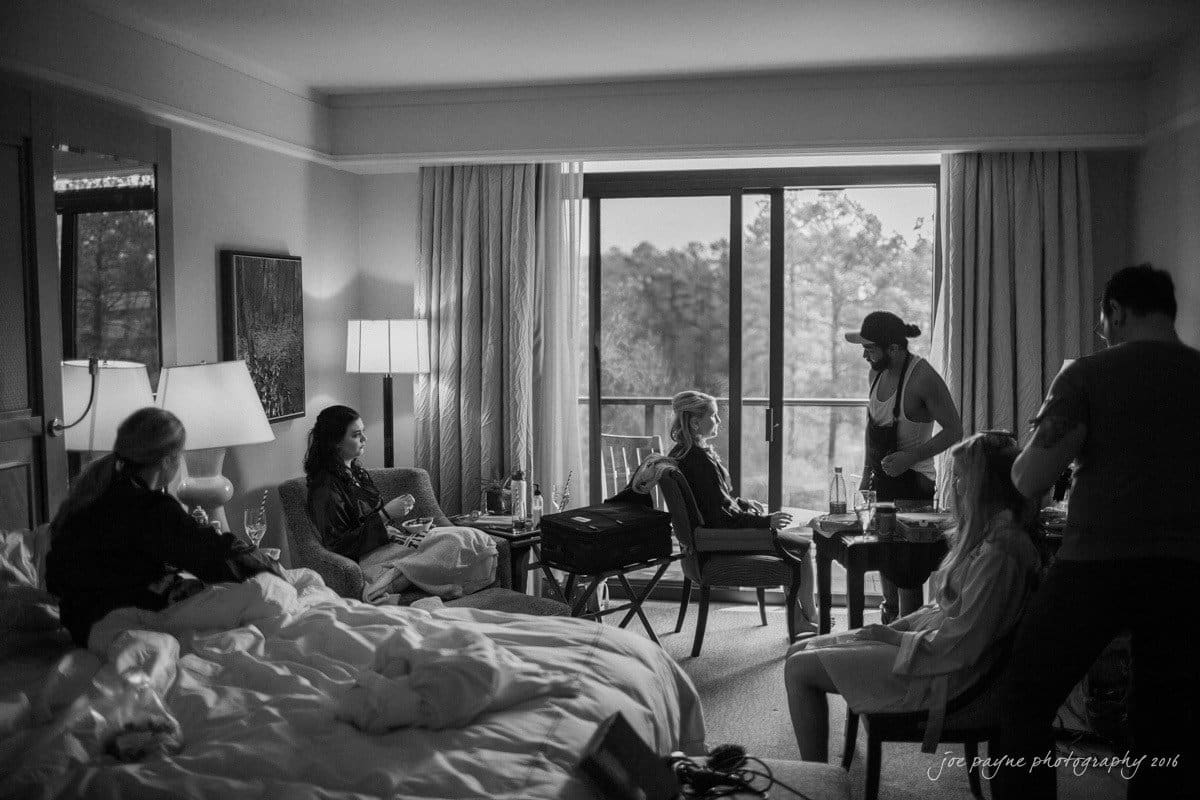 st. mary's chapel raleigh wedding photography - jackie & rob A black and white photo of a hotel room where five people are gathered. Some are sitting on chairs, others on the bed, and one is styling a womans hair. A window in the background offers a view of trees.