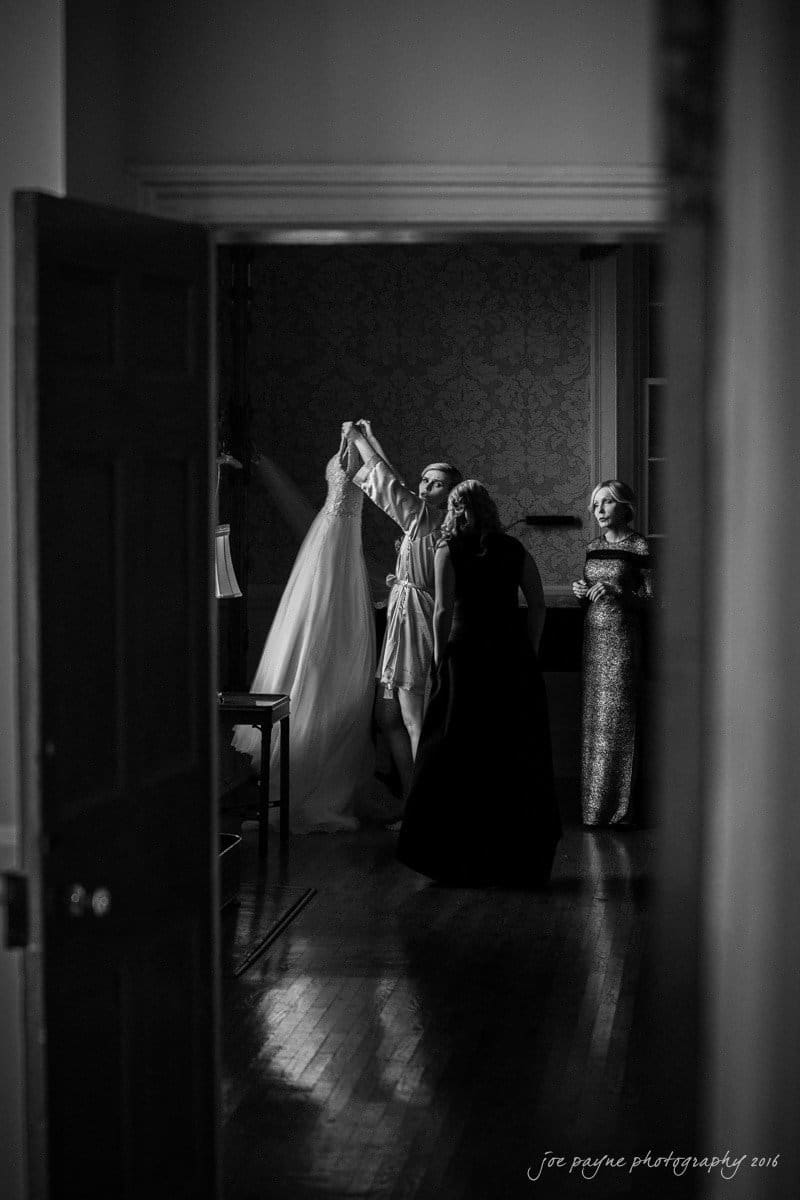 st. mary's chapel raleigh wedding photography - jackie & rob A black and white photo of three women in a dimly lit room. One holds up a wedding dress, while another stands nearby. A third woman in a long dress observes. Theyre surrounded by elegant wallpaper and wooden flooring.