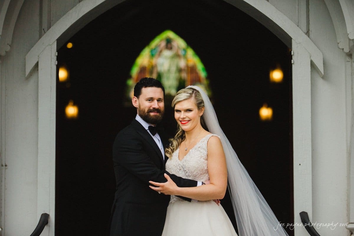 st. mary's chapel raleigh wedding photography - jackie & rob A couple in wedding attire stands smiling in front of a church entrance. The groom wears a black tuxedo, and the bride wears a white gown with a veil. Inside, stained glass windows and warm lights are visible.