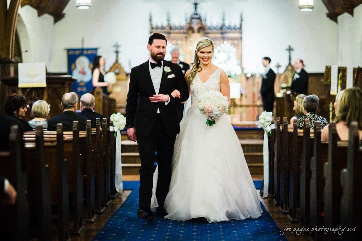 st. mary's chapel raleigh wedding photography - jackie & rob A bride in a white gown and a groom in a black tuxedo smile as they walk down the aisle of a church. The aisle is adorned with white flowers, and guests are seated on wooden pews. An ornate altar is visible in the background.