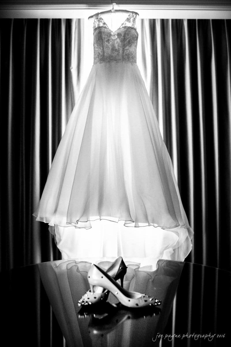 st. mary's chapel raleigh wedding photography - jackie & rob A black and white image of an elegant wedding dress hanging in front of a window with curtains. Below, a pair of embellished high-heeled shoes rests on a reflective surface.