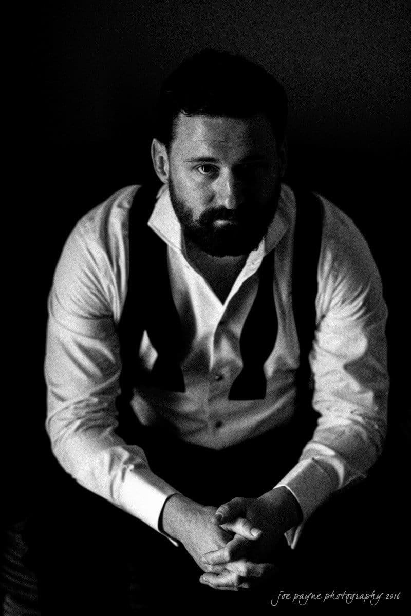 st. mary's chapel raleigh wedding photography - jackie & rob A man with a beard sits in a dimly lit room, wearing a white shirt and a loose, untied bow tie. His hands are clasped together, and he gazes pensively towards the camera. The photo is in black and white.