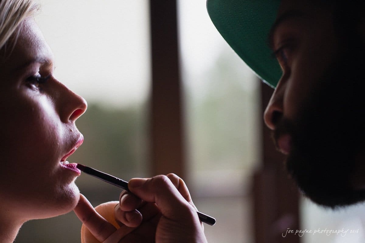 st. mary's chapel raleigh wedding photography - jackie & rob A makeup artist applies lipstick to a persons lips with a brush. The scene is softly lit, creating a focused and intimate atmosphere. Both individuals are partially visible, with the artist concentrated on their work.