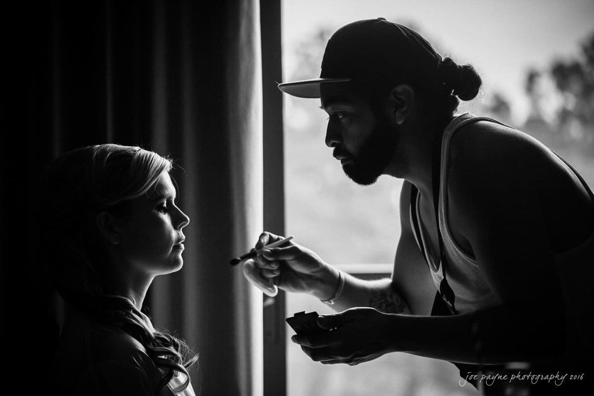 st. mary's chapel raleigh wedding photography - jackie & rob A makeup artist applies cosmetics to a womans face in front of a window. The scene is in black and white, highlighting the focused expressions and the soft natural light filtering through the window.